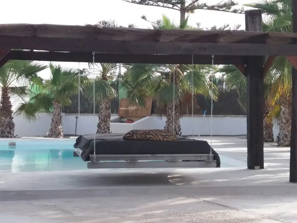 Cama colgante bajo una pérgola de madera oscura junto al agua de una piscina, con palmeras y un muro blanco al fondo