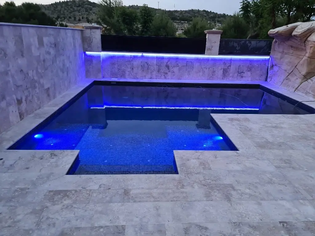Piscina con luz LED azul en el muro de piedra y focos azules bajo el agua, con borde de travertino claro