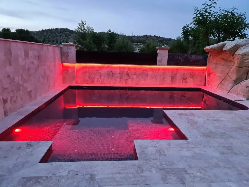 Piscina con luz LED roja en el muro de piedra y focos rojos bajo el agua, con borde de travertino claro