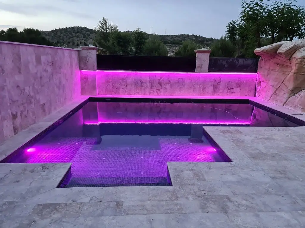 Piscina con luz LED rosa y violeta en el muro de piedra y bajo el agua, con borde de travertino claro