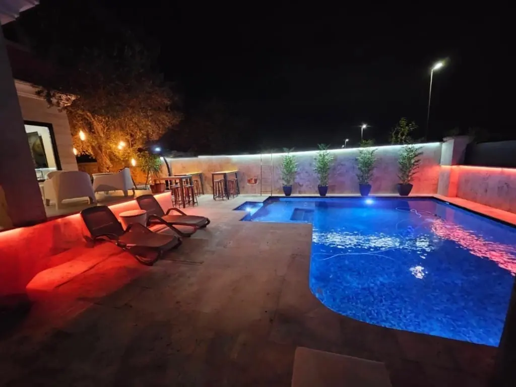 Terraza de noche con tumbonas, muro iluminado en rojo y una piscina de agua azul intenso con spa al fondo