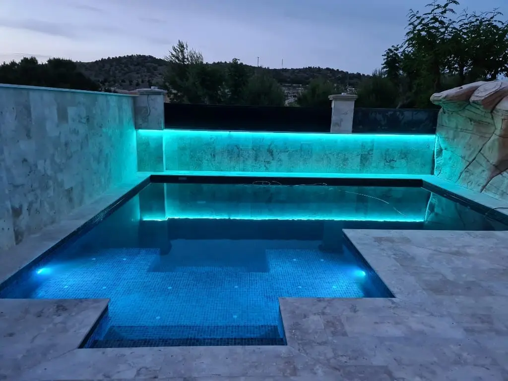 Piscina con luz LED turquesa en el muro de piedra y en el agua, con borde de travertino claro