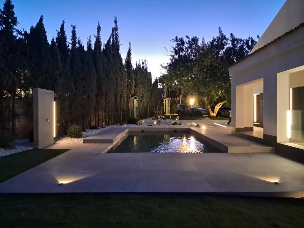 Jardín al anochecer con un espejo de agua iluminado, focos empotrados en el suelo, casa blanca moderna y cipreses