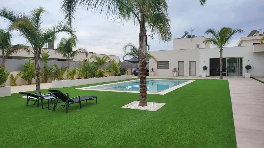 Jardín de césped artificial con una piscina rectangular alargada, dos tumbonas, palmeras y una casa blanca moderna