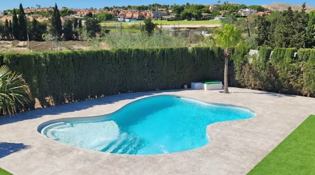 Piscina de forma libre de agua turquesa sobre pavimento impreso gris claro, con césped artificial y seto alto al fondo