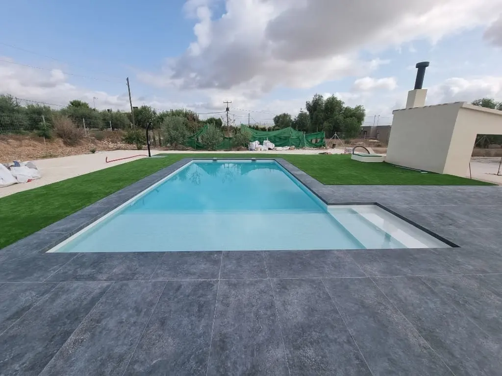 Piscina alargada de agua clara con borde de pavimento gris oscuro, césped y una barbacoa de obra blanca al fondo