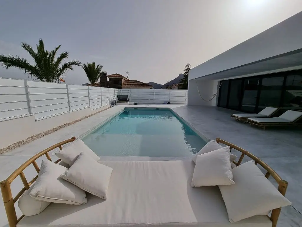 Piscina alargada junto a una casa moderna blanca, con una cama balinesa de cojines blancos en primer plano