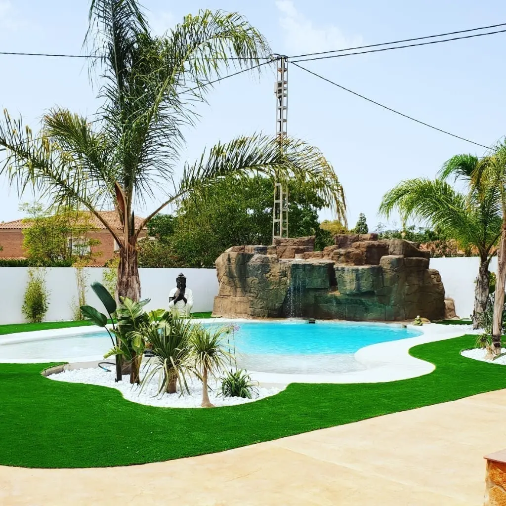 Piscina con cascada de roca, palmeras y césped con piedra blanca decorativa, bajo un cielo despejado