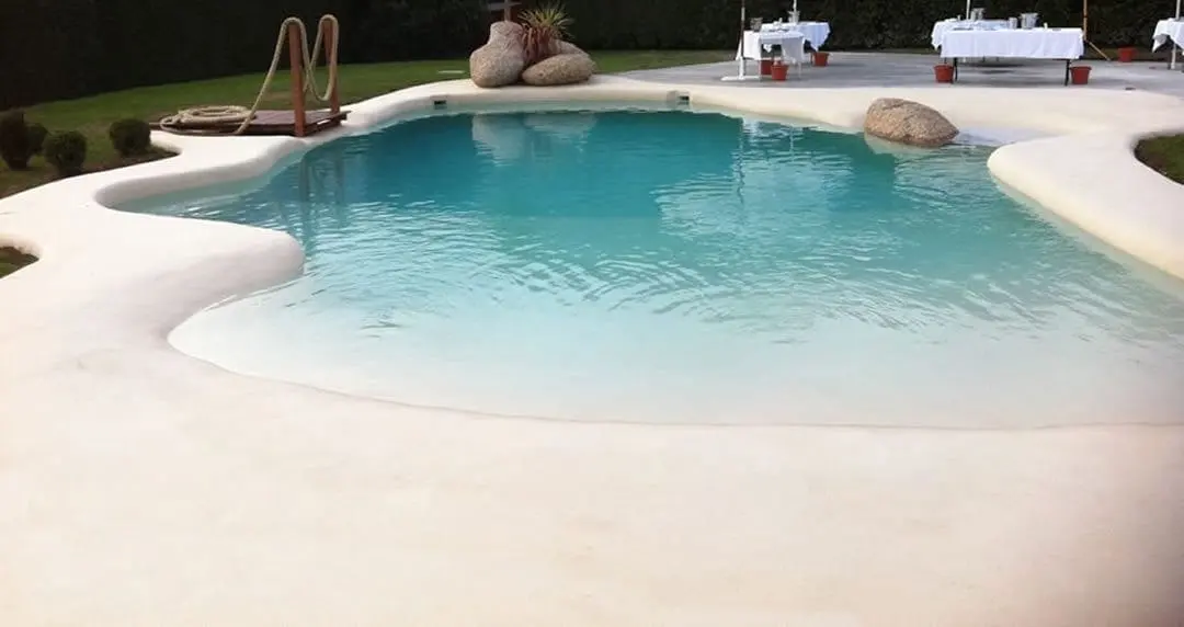 Piscina de arena de forma libre con borde de arena clara y mesas con mantel preparadas al fondo