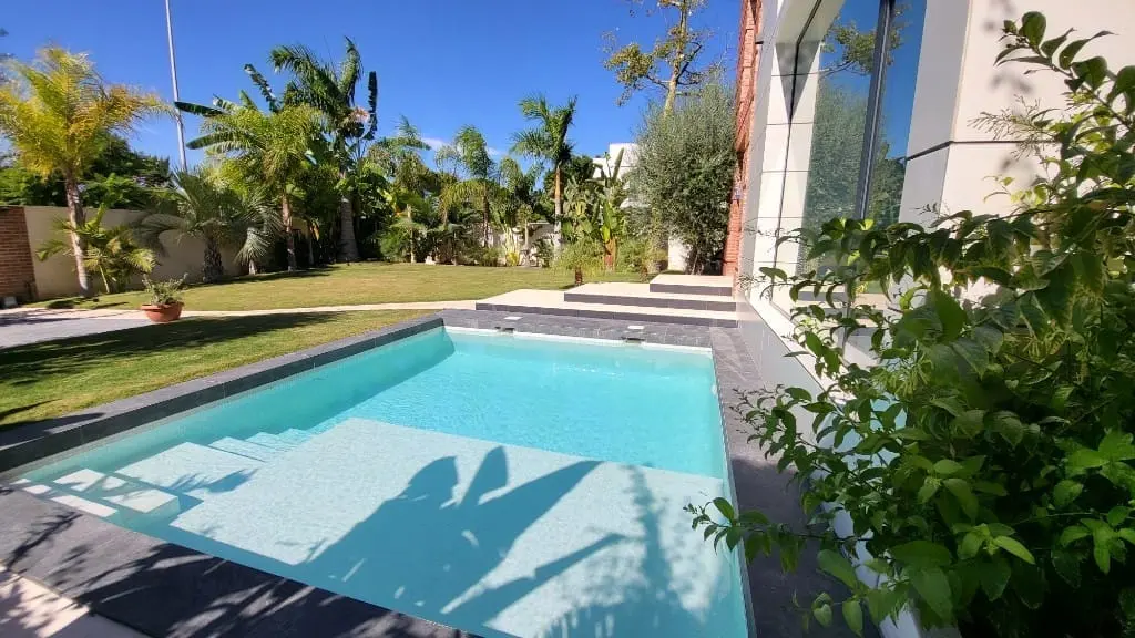 Piscina de agua clara con escalones de acceso, césped, palmeras y vegetación junto a una casa moderna