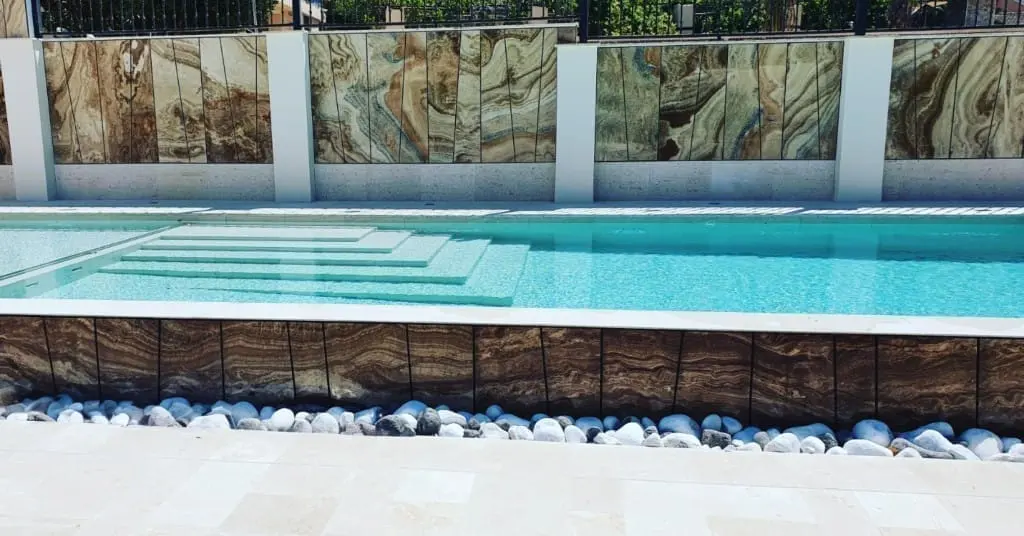 Piscina con escalones sumergidos y un muro decorativo de piedra con vetas verdes y marrones, con canto rodado blanco