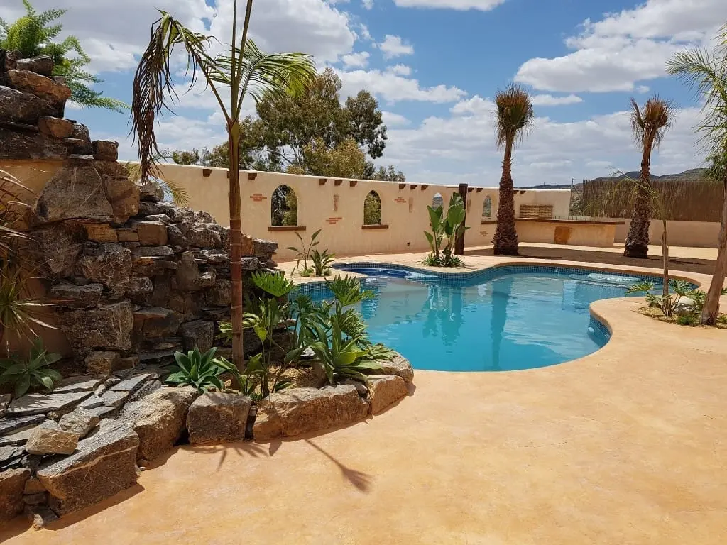 Piscina de forma libre con cascada de roca natural, solárium color arena y palmeras junto a un muro con arcos