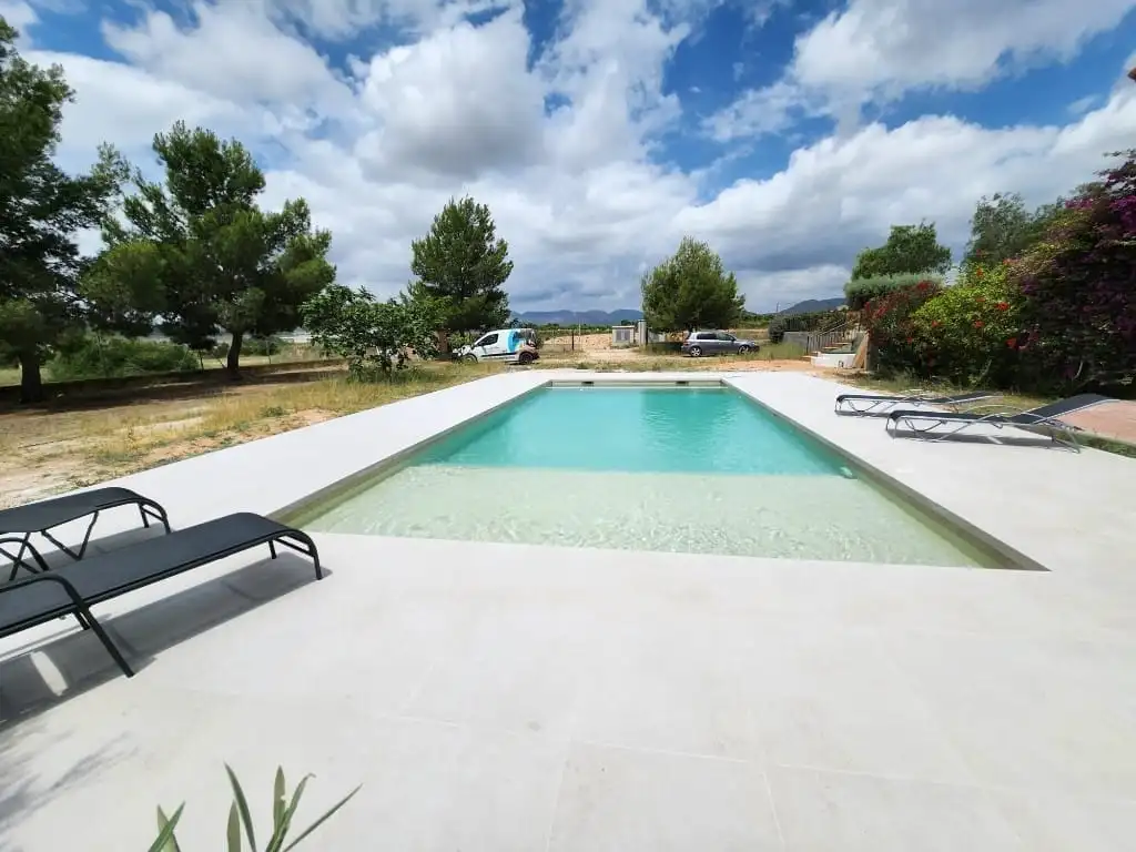 Piscina alargada de agua turquesa con zona de playa y tumbonas, con árboles y campo al fondo