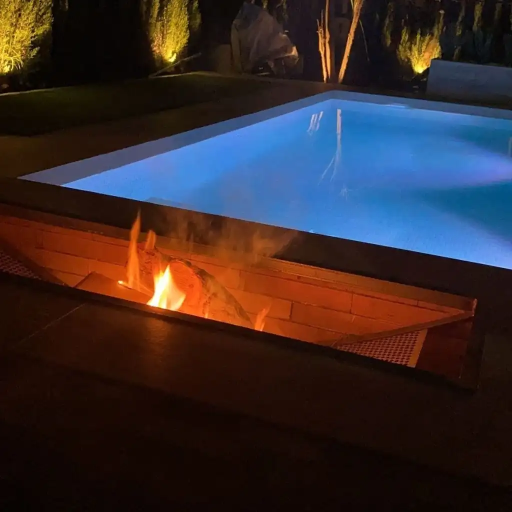 Piscina iluminada en azul por la noche con un fuego decorativo en primer plano