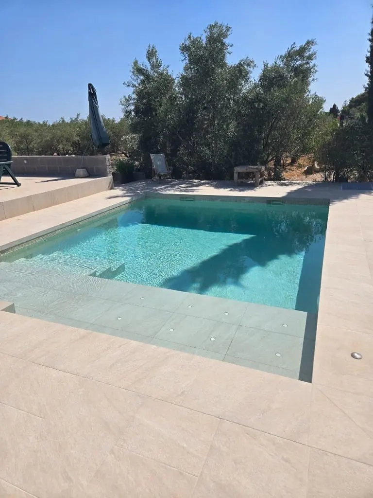 Piscina pequeña de agua turquesa con escalones y pavimento beige, con olivos detrás
