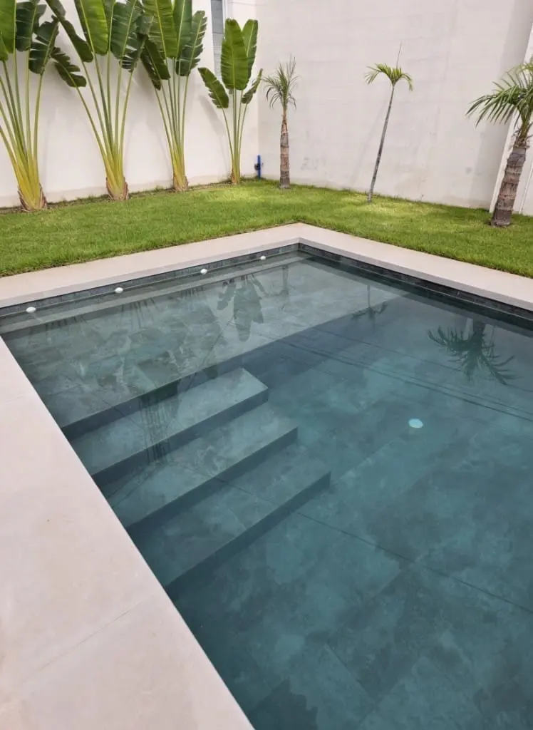 Piscina con escalones revestida de porcelánico gris, junto a un muro blanco con plantas y césped