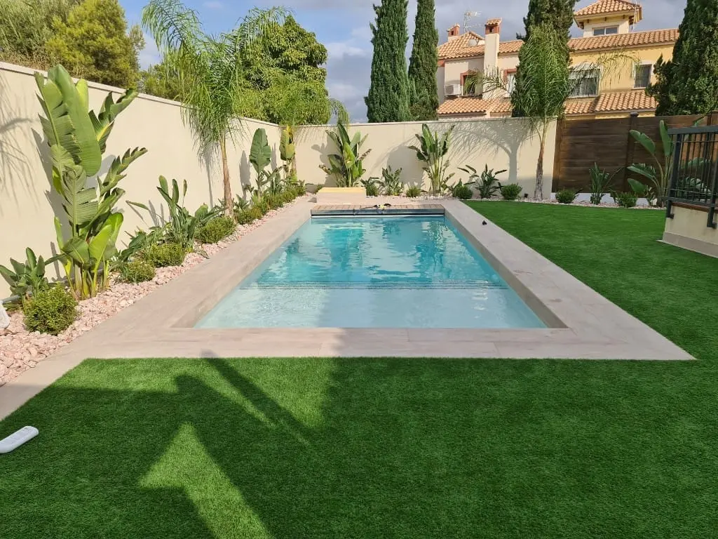 Piscina rectangular de agua clara con borde de piedra beige, rodeada de césped artificial y plantas tropicales