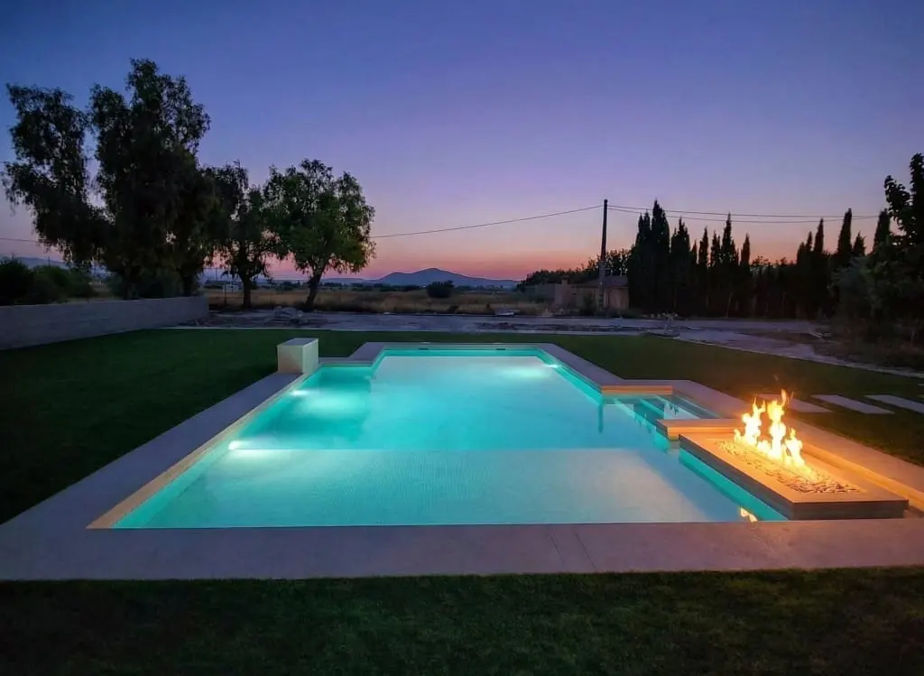 Piscina rectangular iluminada al atardecer con un fuego decorativo en el borde, césped y olivos