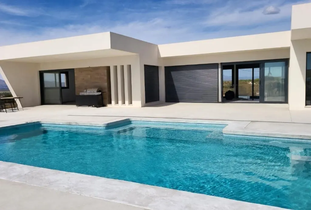 Piscina de agua turquesa junto a una casa moderna blanca de una planta con pavimento claro