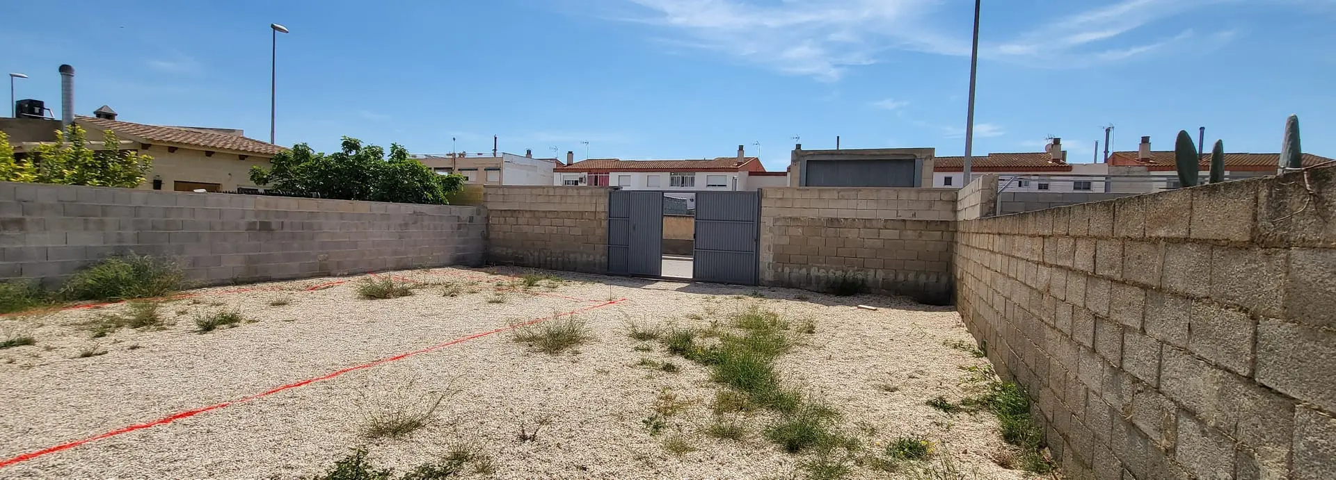 Antes: solar de grava rodeado de un muro de bloques, con puerta metálica y viviendas al fondo, sin piscina