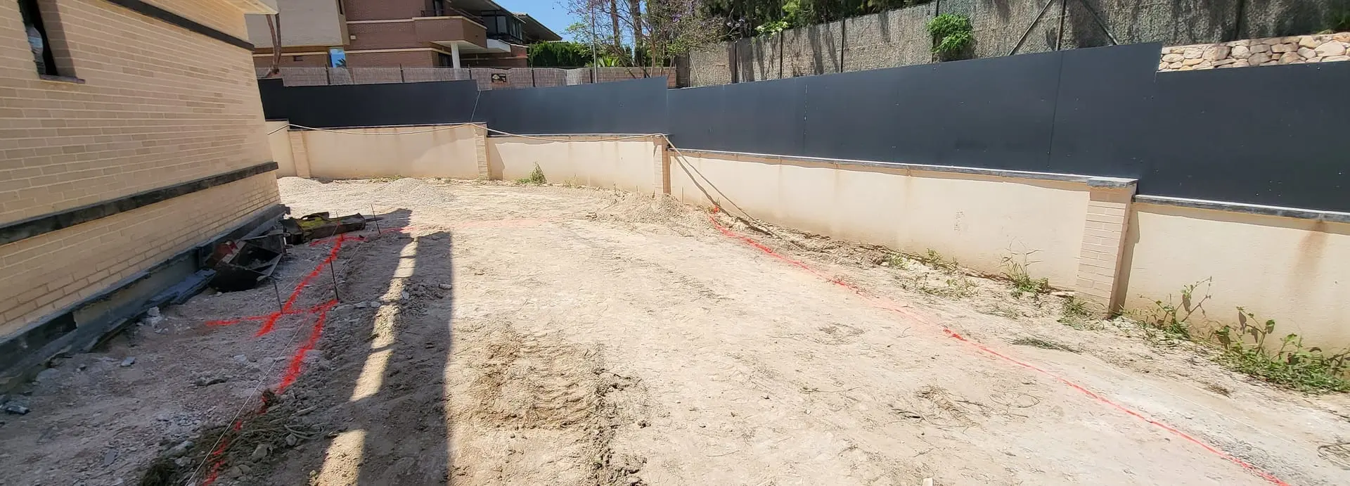 Antes: terreno de tierra con marcas rojas de replanteo junto a una pared de ladrillo y un muro oscuro