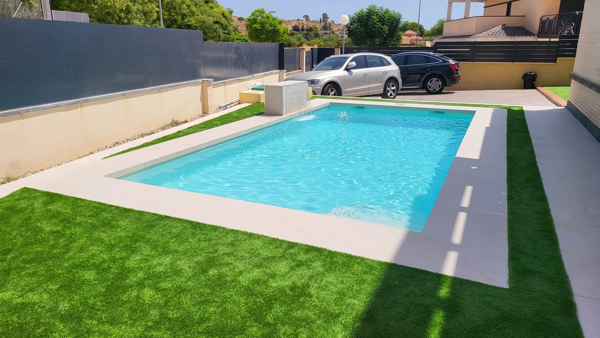 Después: piscina rectangular con borde claro y césped artificial en el patio de una vivienda, con un coche aparcado al fondo