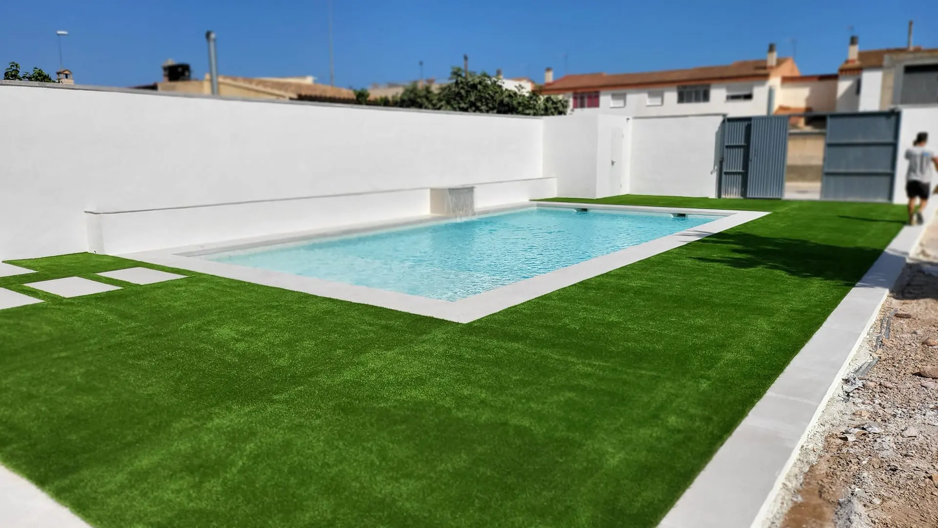 Después: piscina rectangular con borde blanco, césped artificial y losas de paso junto a un muro blanco