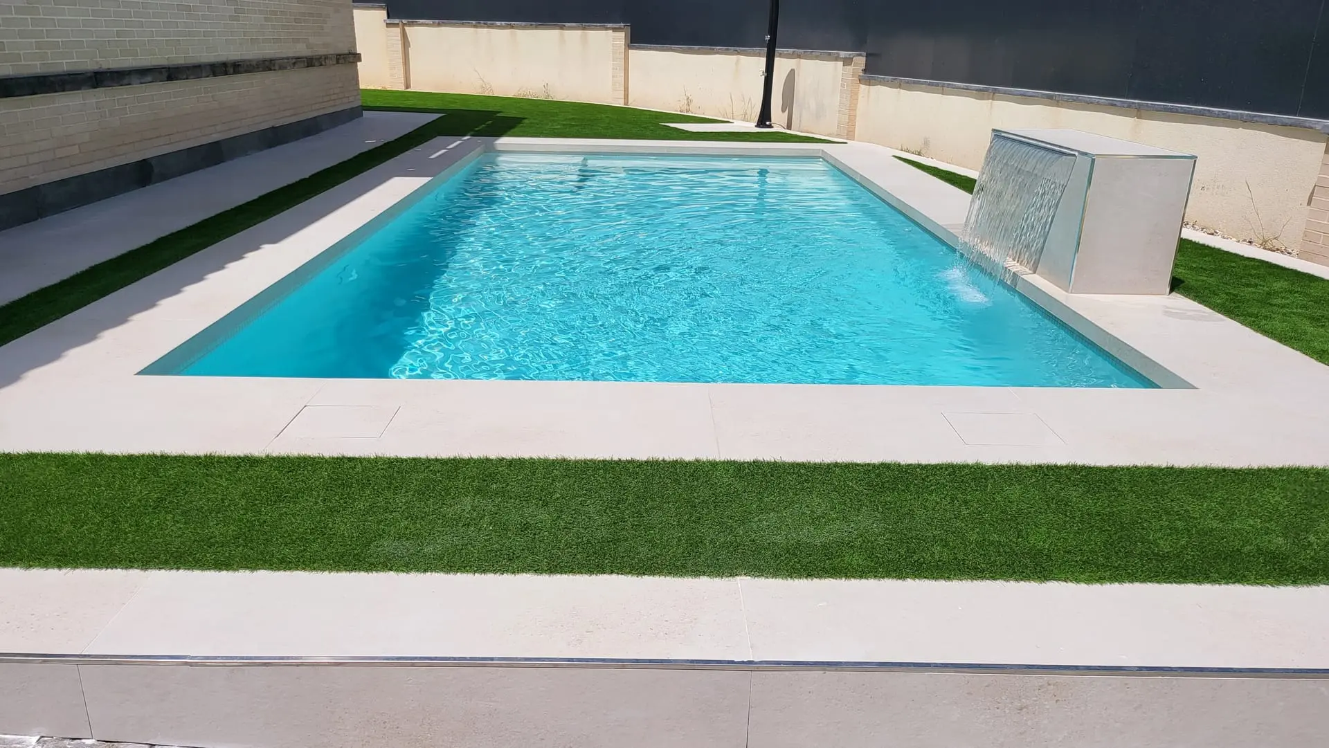 Después: piscina rectangular de agua turquesa con cascada de lámina, borde claro y césped artificial
