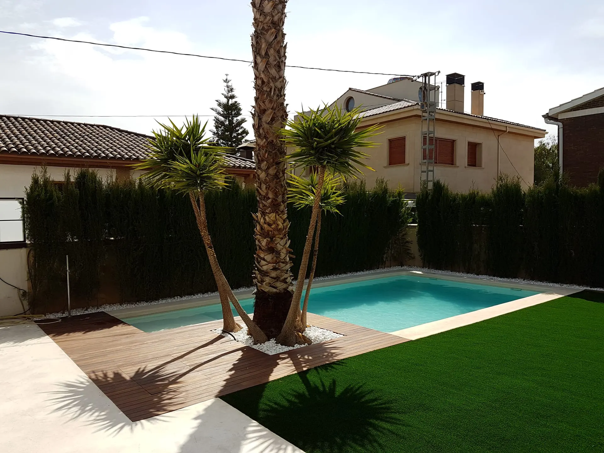Después: piscina de agua turquesa con tarima de madera, palmera, césped artificial y seto de cipreses